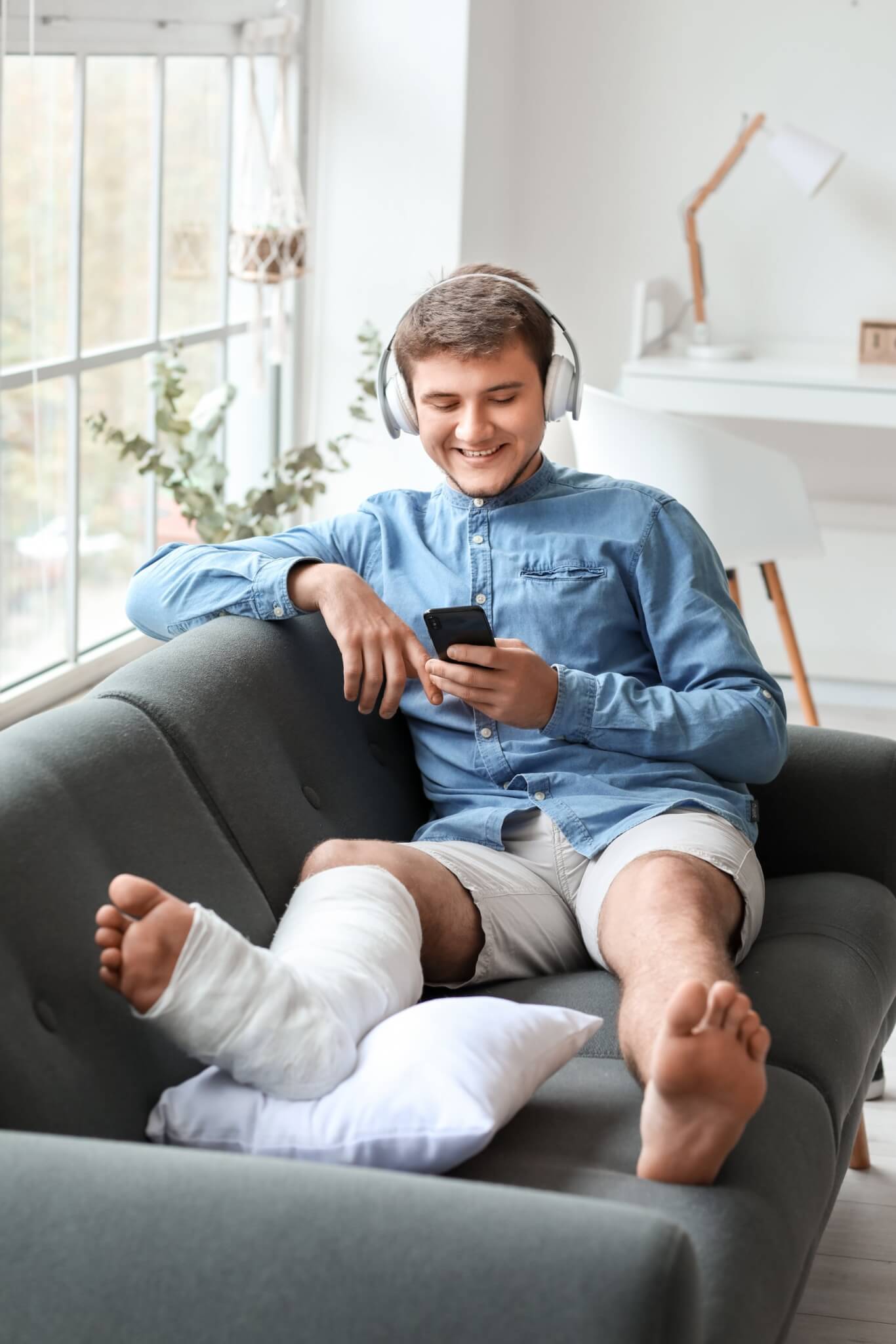 young adult man with broken leg sitting on couch at home listening to music through headphones